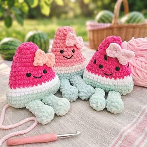 Può includere: Tre peluche all'uncinetto a forma di fette di anguria, ciascuno con una parte superiore rosa, una centrale bianca e una base verde chiaro. Ogni peluche ha occhi neri, un sorriso ricamato e un piccolo fiocco rosa chiaro nell'angolo superiore.