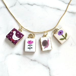 Könnte beinhalten: Eine goldfarbene Halskette mit vier rechteckigen Anhängern. Die Anhänger zeigen Illustrationen einer weißen Gans auf violettem Grund, einer rosa Blume, einer braunen Schnecke und einer violetten Blume auf cremefarbenem Grund, alle mit Goldringen.