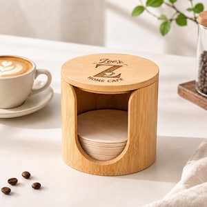 Może przedstawiać: Cylindryczny stojak na podstawki z jasnobrązowego bambusa ze zdejmowaną pokrywką, na której wygrawerowano napis "Zoe's HOME CAFE" oraz dużą literę Z z ikoną małej filiżanki kawy. Wewnątrz stojaka znajduje się stos okrągłych podstawek pod kubki.