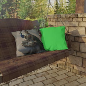 May include: A wooden porch swing with two decorative pillows. One pillow features a Buddha statue with a peacock, and the other is solid green. The swing has a brown plaid blanket and is hanging near a brick wall.