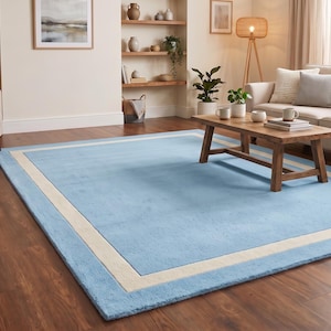May include: A rectangular area rug featuring a solid light blue center field surrounded by a wide, cream-colored border. The rug has a low-pile texture and is positioned flat on a dark wood floor, covering a large portion of the living room space.