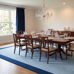 May include: A formal dining room featuring a long wooden table with eight chairs, each with cream-colored upholstered seats. A light blue area rug with a dark blue border sits on the hardwood floor. Navy blue curtains frame the window.