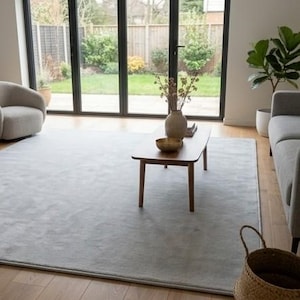 May include: A large, rectangular, light gray area rug covers the wooden floor in a living room. The rug features a solid, uniform color and a soft texture, positioned centrally beneath a wooden coffee table, a gray sofa, and a gray armchair.