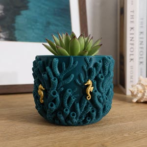 May include: A dark teal cylindrical planter featuring a textured, raised coral reef pattern across its exterior. Two small, gold-colored seahorse figures are attached to the sides of the planter. A green succulent plant is potted inside the container.