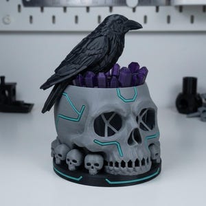 May include: A gray skull-shaped decorative object with a black raven perched on top. The skull has teal accents and is filled with purple crystals. Small skulls adorn the base, creating a gothic and unique aesthetic.