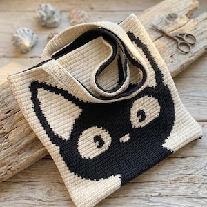 Puede incluir: Un bolso tote cuadrado de ganchillo con un diseño de cara de gato en negro y crema. El bolso está construido con un patrón de punto denso y texturizado e incluye dos asas de ganchillo a juego. El gráfico del gato cubre el frente con ojos grandes.