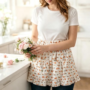 May include: A white short-sleeved t-shirt paired with a white waist apron featuring a repeating orange floral pattern. The apron includes two front pockets and a ruffled hemline, worn over dark blue jeans while holding a small bouquet of pink roses.