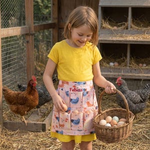 May include: A light pink egg collecting apron featuring a repeating pattern of blue and orange chickens, with the name Felicia embroidered in red thread. The apron has multiple pockets for holding eggs and is worn over a yellow t-shirt.