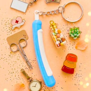 May include: A collection of miniature medical-themed accessories, including a blue and white toothbrush, a nurse's hat charm, a pill bottle, a watch, scissors, and a heart-shaped pin. The items are arranged on a peach-colored surface with gold glitter.