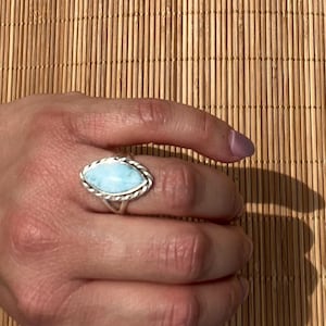 May include: A silver ring with a light blue, oval-shaped stone. The ring has a twisted rope design around the stone. The ring is on a hand, with a bamboo mat in the background.