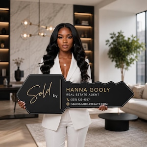 May include: A large, black, key-shaped sign with gold lettering that reads Sold by HANNA GOOLY REAL ESTATE AGENT, a phone icon with (555) 123-4567, and a globe icon with HANNAGOOLYREALTY.COM. The sign is held by a person in a white blazer.