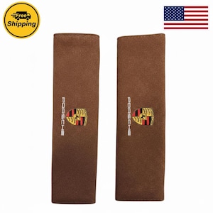 May include: A pair of rectangular brown seat belt covers featuring the white text PORSCHE and the colorful Porsche crest logo centered on each piece. The covers have a soft, textured surface and are presented side by side against a plain background.
