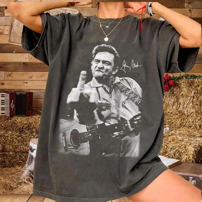 May include: Dark gray t-shirt featuring a black and white image of Johnny Cash playing a guitar and giving the middle finger. The shirt has a vintage, worn look. The signature "Johnny Cash" is printed on the shirt.