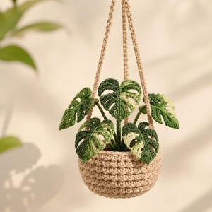 Pode incluir: Uma planta suspensa em crochê com um vaso de fio bege e cordas longas a condizer. A planta apresenta várias folhas de monstera variegadas em tons de verde escuro e creme claro, trabalhadas com pontos detalhados que imitam a folhagem.