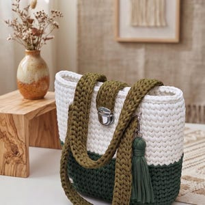 May include: A two-toned crochet tote bag featuring a white upper section and a dark green lower section. The bag includes two olive green braided shoulder straps, a silver-toned metal clasp closure, and a matching dark green fabric tassel.