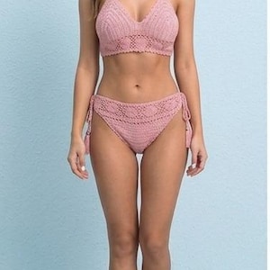 May include: A two-piece crochet bikini set in a soft pink hue. The top is a halter style, and the bottoms are high-waisted with tie details. The set is made of a textured, open-weave material, perfect for swimwear.