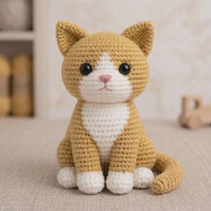 May include: A crocheted cat figurine in shades of tan and white. The handmade cat has large black eyes, a pink nose, and white paws. The cat is sitting upright with its tail curled to the side. The cat is a handmade item.