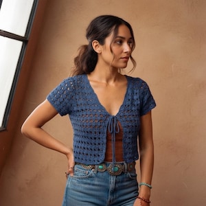 Può includere: Un top all'uncinetto blu, a maniche corte e aperto sul davanti, con scollo a V e chiusura con lacci. Il top presenta un delicato motivo traforato. La modella indossa jeans blu e una cintura turchese. Lo sfondo è di un colore neutro e caldo.