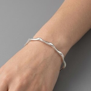 Peut inclure: Un bracelet argenté avec un bord ondulé est porté sur un poignet. Le bracelet a un design fluide et organique. Le fond est gris neutre.