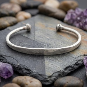 Könnte beinhalten: Ein silberfarbenes Armband in Manschettenform mit einer gehämmerten, strukturierten Oberfläche und abgerundeten, knopfartigen Enden. Das Armband hat ein offenes Design zur Anpassung und eine matte, metallische Oberfläche.