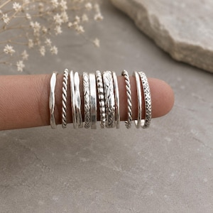 May include: A collection of eleven silver-toned rings arranged in a row on a finger. The rings feature various designs including smooth bands, twisted rope textures, beaded patterns, engraved floral motifs, and cross-hatch geometric detailing.