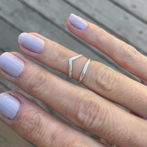 May include: A hand with light purple nail polish, showcasing two silver rings. One ring is a plain band, the other a V-shaped design. The rings are worn on the middle finger.