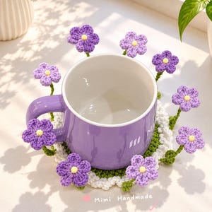 Puede incluir: Una taza de cerámica morada descansa sobre un posavasos redondo de ganchillo blanco y verde, decorado con varias flores pequeñas de lana morada con centros amarillos. El texto "Mimi Handmade" con un corazón rosa es visible en la parte inferior.