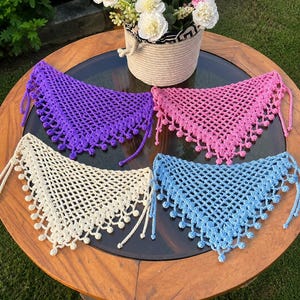 May include: Four crocheted triangle scarves in purple, pink, cream, and light blue are displayed on a round wooden table. Each scarf has a delicate open weave and is trimmed with small, decorative bobbles and ties. A basket of flowers sits in the background.