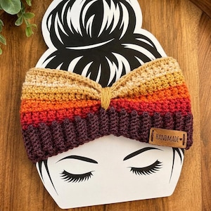 May include: A crochet ear warmer headband featuring a horizontal striped pattern in shades of beige, yellow, orange, red, and dark purple. The center is cinched with a yellow yarn knot and includes a small brown leather tag labeled HANDMADE.