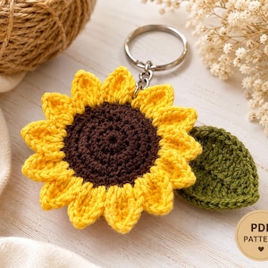 Può includere: Un portachiavi a forma di girasole all'uncinetto con un centro marrone scuro, petali giallo brillante e una singola foglia verde attaccata a un anello in metallo argentato. Un badge circolare beige riporta il testo PDF PATTERN con un cuore.