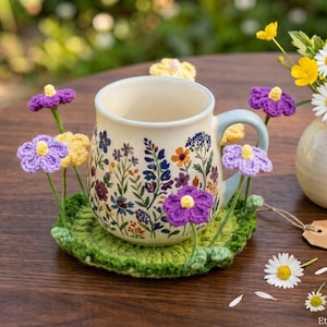 Puede incluir: Una taza de cerámica con un patrón de flores silvestres de colores se asienta sobre un posavasos de ganchillo verde decorado con pequeñas flores de hilo moradas y amarillas. Un pequeño jarrón blanco contiene un ramo de margaritas y ranúnculos amarillos, con una etiqueta que dice The Crochet Meadow - Flower Cozy Set.