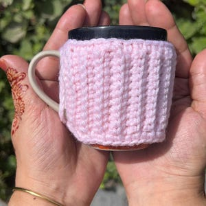 May include: A pink, knitted mug cozy. The cozy is wrapped around a white mug with a black lid. The mug is held in two hands. The cozy is made of a soft, textured yarn. The mug cozy is designed to keep beverages warm.
