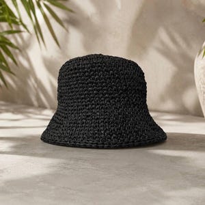 May include: A solid black bucket hat featuring a dense, textured crochet knit construction. The hat has a rounded crown and a downward-sloping brim that encircles the entire head opening, showcasing a consistent, uniform weave pattern throughout.