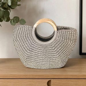 Könnte beinhalten: Eine halbmondförmige Handtasche aus einem strukturierten, hellgrauen Webmaterial. Die Tasche verfügt über einen markanten, kreisförmigen Griff aus hellem Holz, der in die obere Mitte integriert ist und einen festen Halt zum Tragen bietet.