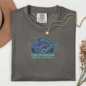 May include: A charcoal gray t-shirt with a Comfort Colors label in the collar. The chest features a circular graphic with the text "FUTURE INNOVATORS," "READY TO CODE & CREATE," "TECH EXPLORATION," and "THE DIGITAL ERA EST. 2026 GLOBAL LAB" with icons of a laptop, VR headset, robot, and rocket.