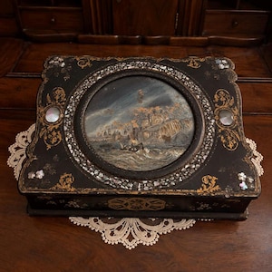 May include: A rectangular black lacquered box featuring a central circular panel with a painted coastal scene of a ship at sea near cliffs. The box is decorated with gold floral filigree, inlaid mother-of-pearl accents, and small flower motifs.