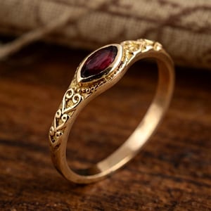 May include: A gold ring featuring a marquise-cut deep red gemstone set in the center. The band is crafted with intricate metalwork patterns along the sides, showcasing a detailed scroll design that extends from the central stone setting.