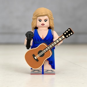 May include: A Lego figurine of a female singer with blonde hair, wearing a blue dress, holding a black microphone and a brown acoustic guitar. The guitar has black tuning pegs and white strings. The figurine has red lipstick.