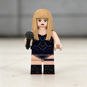 May include: A Lego figurine of a performer with blonde hair and red lipstick, holding a black microphone. The figurine wears a dark blue outfit with a decorative pattern and black boots with white dots.