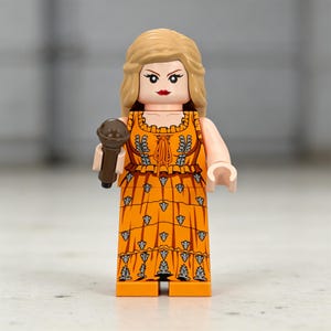 May include: A Lego figurine of a person with blonde hair, wearing an orange dress with a floral pattern, and holding a brown microphone. The figurine has a red lipstick and is standing upright.
