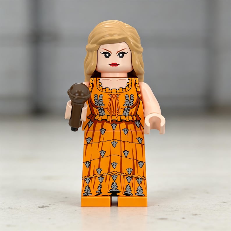 May include: A Lego figurine of a person with blonde hair, wearing an orange dress with a floral pattern, and holding a brown microphone. The figurine has a red lipstick and is standing upright.
