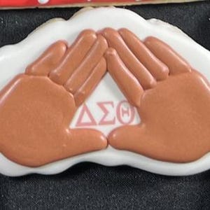 May include: A decorated cookie with a white base and a design featuring two hands in a clasping gesture. The hands are a light brown color, and the cookie includes the Greek letters ΔΣΘ in pink.
