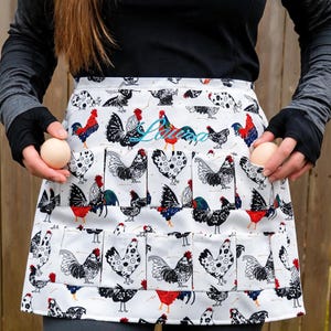 May include: A white waist apron featuring a repeating pattern of black, white, and red roosters and hens. The apron includes multiple pockets for holding eggs and has the name Laura embroidered in blue script across the upper center section.
