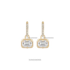 May include: A pair of gold hoop earrings featuring a rectangular emerald-cut diamond pendant suspended from each hoop. The hoops are accented with small pavé-set diamonds. The text OURADIAMONDS is visible near the earrings and at the bottom.