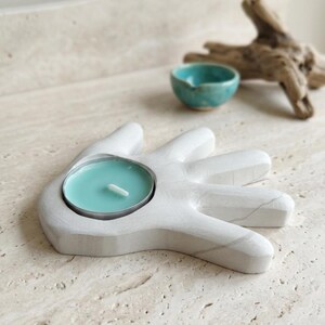 May include: A white hand-shaped candle holder with a smooth, matte finish and subtle grey veining. A round teal-colored tea light candle sits securely in the center of the palm. The object is crafted with five distinct finger-like extensions.