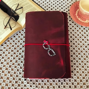 May include: A deep burgundy leather journal secured with a bright red elastic band. A decorative charm shaped like black and white chequered eyeglasses with small red accents is attached to the elastic band, resting on the front cover of the journal.