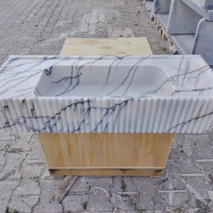 May include: A rectangular white marble sink featuring prominent dark blue veining throughout the stone. The exterior of the basin is designed with a vertical fluted texture, and the top surface includes a flat deck area surrounding the central basin.