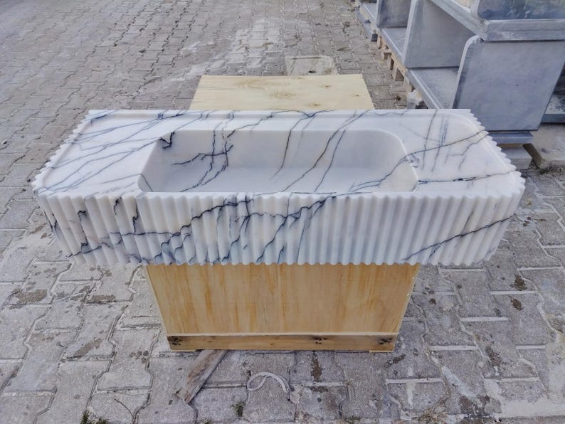 May include: A rectangular white marble sink featuring prominent dark blue veining throughout the stone. The exterior of the basin is designed with a vertical fluted texture, and the top surface includes a flat deck area surrounding the central basin.