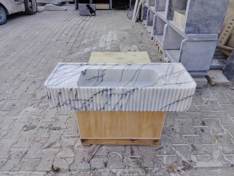 May include: A rectangular white marble sink featuring prominent dark gray veining and a fluted, vertically ribbed exterior design. The basin is integrated into a flat countertop surface, showcasing a polished stone texture and a clean, linear aesthetic.