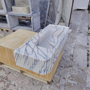 May include: A rectangular white marble sink featuring prominent dark blue veining and a fluted, vertically ribbed exterior design. The basin has a smooth interior surface with a central drain hole, resting on a light-colored wooden support structure.